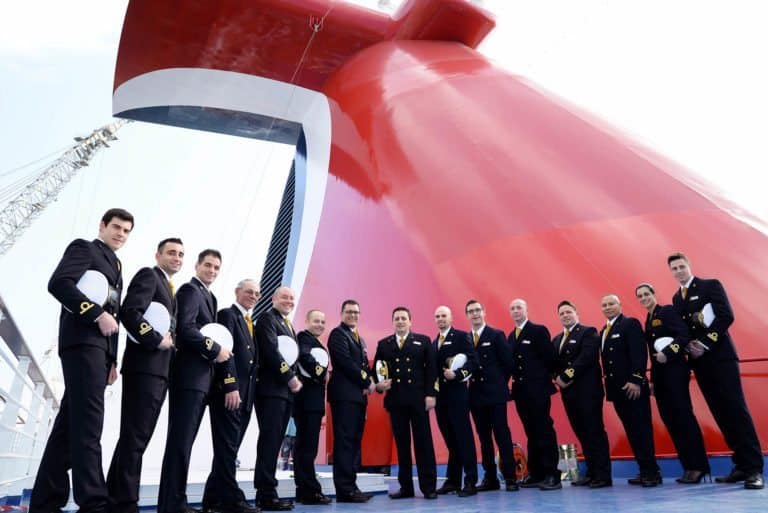 A group of 14 individuals in uniform, likely Carnival cruise ship crew members, stand in a line in front of a large red ship funnel. They are holding their hats and smiling. The background shows part of the new cruise ship and a crane.