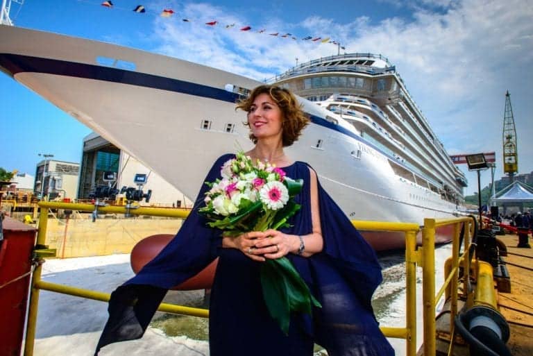 A woman in a dark blue dress holding a bouquet of flowers stands in front of a large Viking ocean ship docked at a shipyard. The sky is blue with scattered clouds, and various structures and equipment are visible around the area.