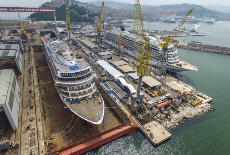 An aerial view of a bustling shipyard with two large Viking cruise ships docked. Yellow cranes are positioned around the vessels, and work areas and equipment are visible on the docks. Several smaller boats and buildings float in the background.