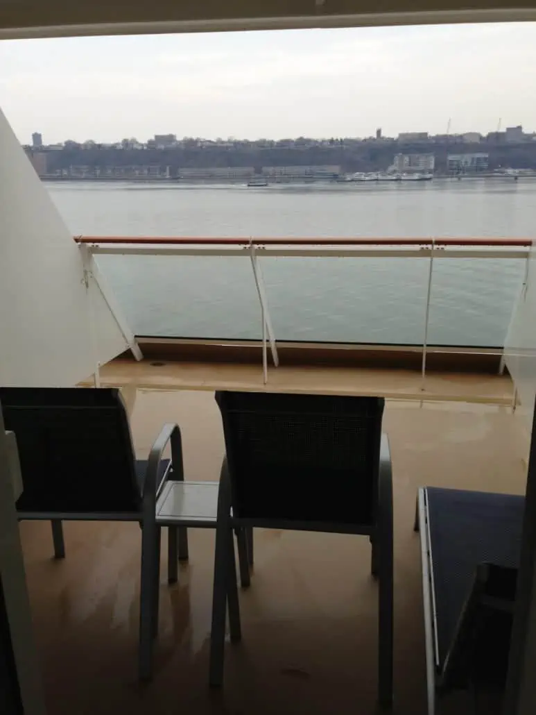         View from an aft balcony cabin overlooking a calm body of water on a cloudy day. Two chairs and a small table are placed on the deck. A distant shoreline with buildings and trees is visible across the water, offering both pros and cons for cruising enthusiasts seeking serenity.