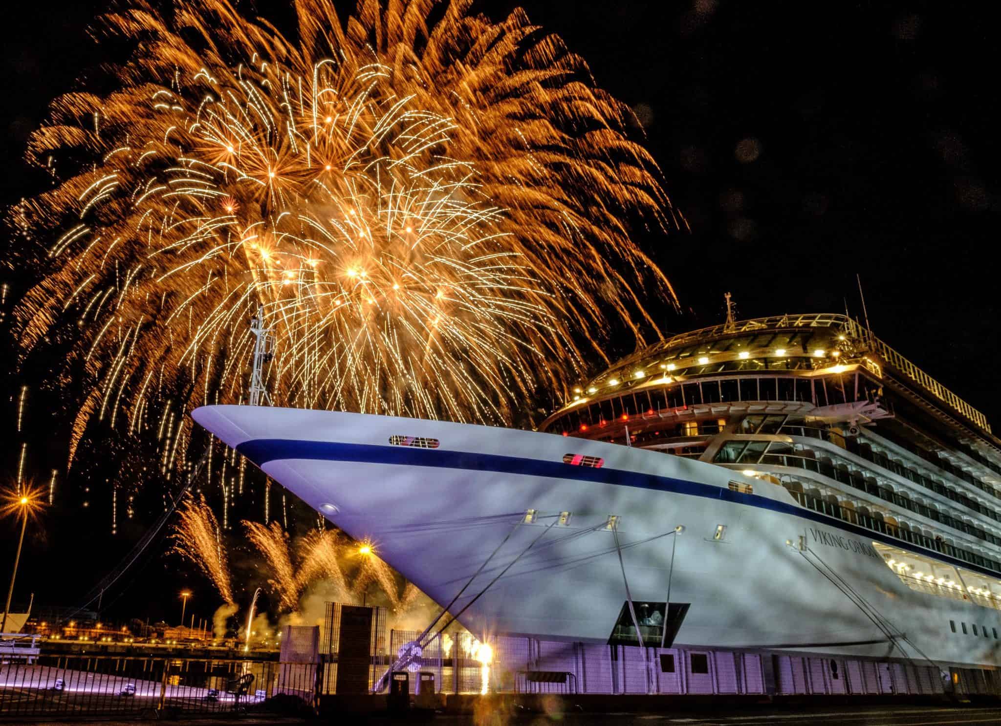 A large Viking Ocean Ship is docked at night with vibrant fireworks bursting in the sky above it. The ship's brightly lit windows and the colorful fireworks create a festive atmosphere reminiscent of a summer evening in Italy.