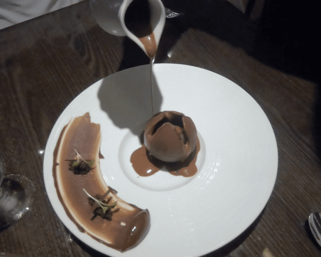 A dessert on a white plate is being drizzled with brown sauce at Fahrenheit 555 Steakhouse. The dish includes a curved piece of chocolate with decorative streaks on the left and a round chocolate confection at the center, set against the backdrop of a dark wooden table.
