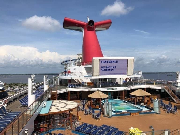 A cruise ship deck with a central pool area, several lounge chairs, and two thatched umbrellas offers a perfect retreat in the outdoor areas. The Carnival Sunshine's iconic red and white funnel stands proudly in the background. A large screen displays a message: "A Fond Farewell! Safe travels and ciao for now!