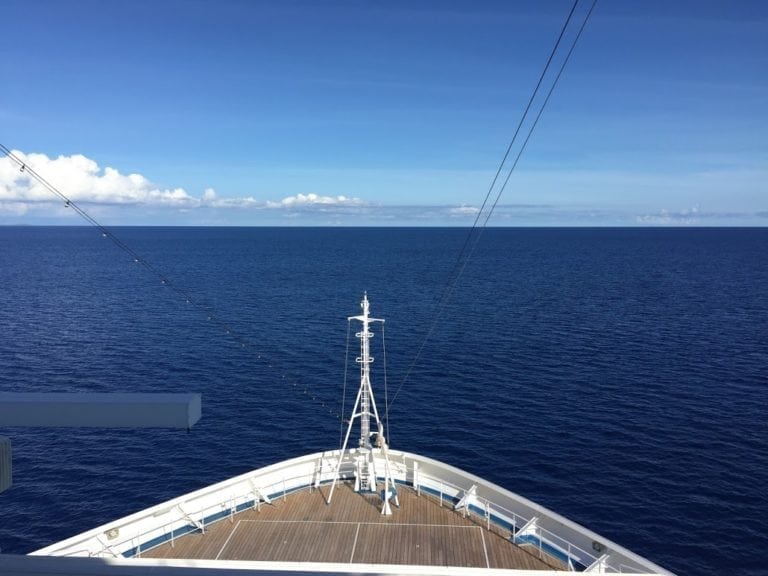 The image shows the bow of a large ship, possibly the Carnival Sunshine, cruising on a calm, deep blue ocean under a clear blue sky. The ship's deck is visible, stretching out towards an expanse of open water with a few clouds on the distant horizon in these beautiful outdoor areas.