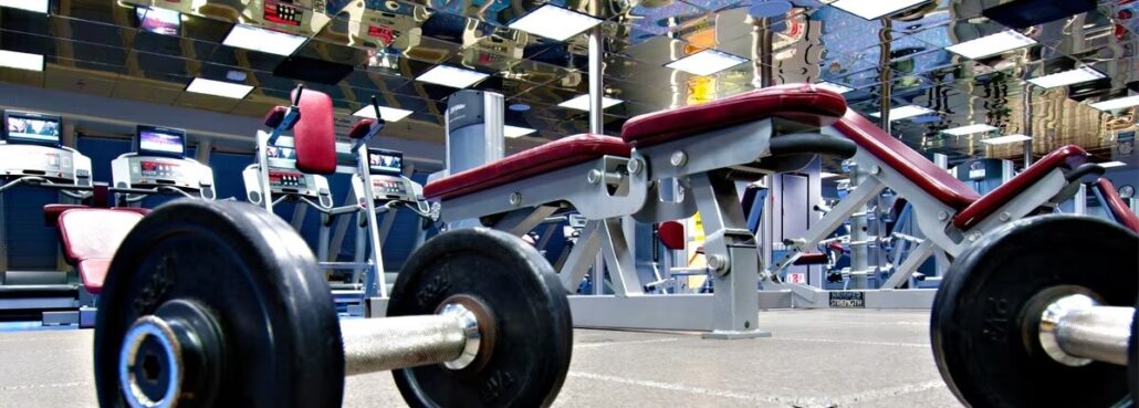 A gym interior on the Carnival Horizon features barbells, weight benches, and treadmills. The floor is carpeted, and the ceiling is lined with reflective tiles. The gym equipment is arranged neatly, with a focus on weightlifting and cardio machines for your cruise fitness routine.