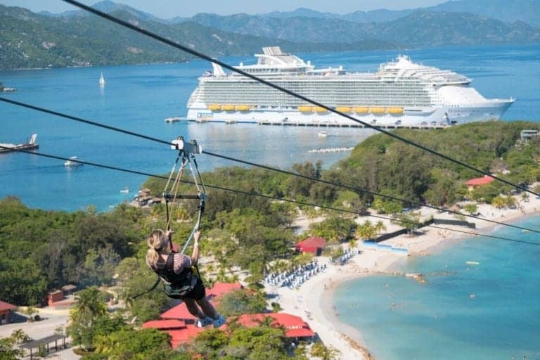 A person rides a zip line above a beach with clear blue water and green foliage. Below, the small resort area boasts red-roofed buildings, white beach chairs, and umbrellas. In the background, a Royal Caribbean cruise ship is docked near the shore, adding to the at-sea adventure vibe.