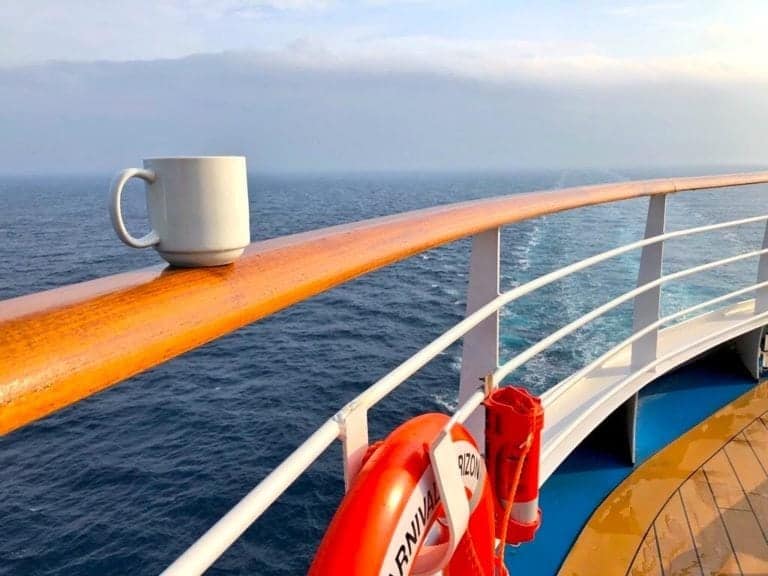 A white coffee mug sits on a wooden handrail of a ship at sea. Below, a lifebuoy and a red emergency box are attached to the railing. The ship is on the ocean, with water and a cloudy sky visible in the background.