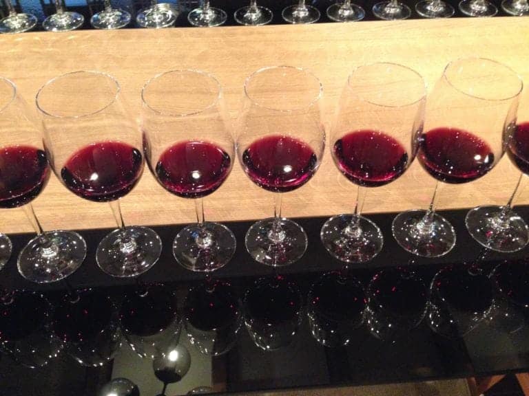 Eight glasses of red wine are lined up in a row on a black reflective surface, perfect for wine lovers. The wine has a dark red hue and is filled to the same level in each glass. A wooden background, reminiscent of the cozy cabins on Norwegian Cruises, is visible behind the row of glasses.