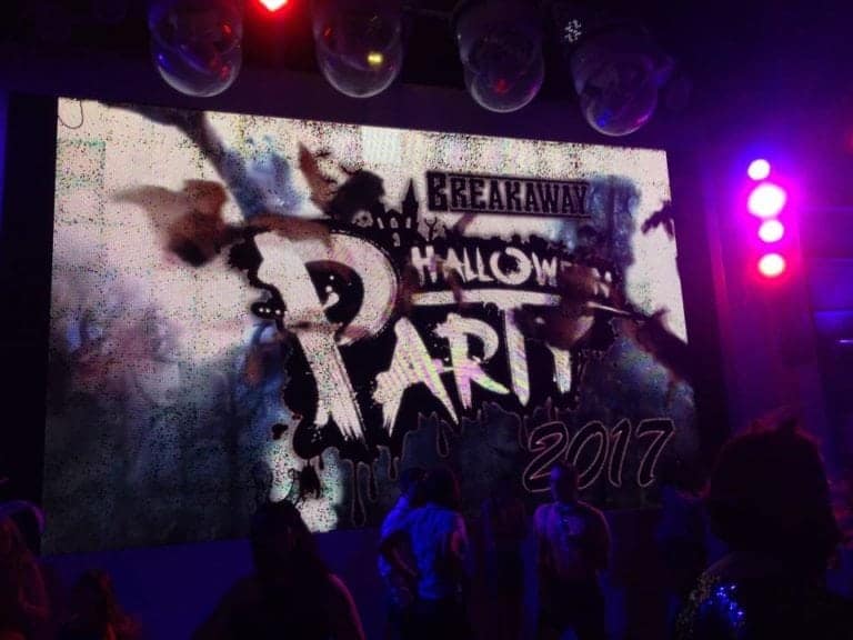 A large screen displays the text "Breakaway Halloween Party 2017" with a graffiti-like design. The screen is surrounded by purple and red lighting, casting an eerie glow over costumed partygoers enjoying various Halloween activities, creating an atmosphere reminiscent of a spooky Halloween cruise experience.