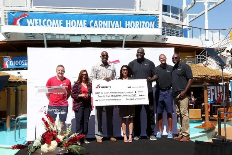 A group of seven people stand on a stage outdoors with a large check. The check is addressed to St. Jude Children's Research Hospital for $25,000. A "Welcome Home Carnival Horizon" sign is visible in the background, along with a ship and floral arrangements, marking the ship's new home port celebration.