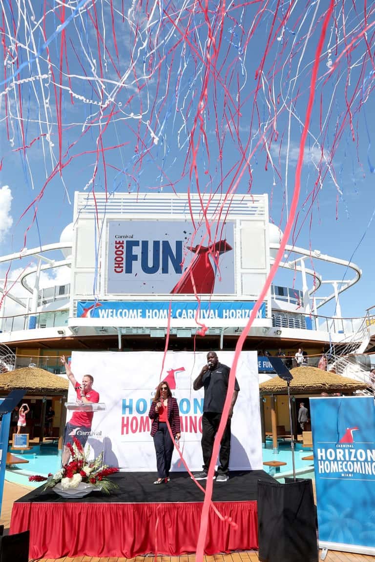 A man and a woman stand on a stage on the Carnival Horizon, speaking into microphones. Behind them, a large sign reads "Choose Fun" and "Welcome Home Carnival Horizon." Red and white streamers are in the air above them. The stage has a banner that reads "Horizon Homecoming," celebrating its new home port.