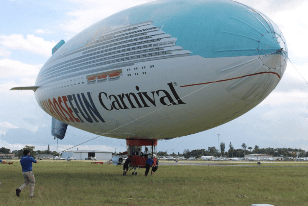 A large blimp, designed to resemble a Carnival cruise ship with the phrase "Choose Fun" written on the side, is tethered to the ground. Several people are seen holding guide ropes to stabilize the blimp, which prominently features the logo of Carnival Corporation. A small platform is suspended beneath it.