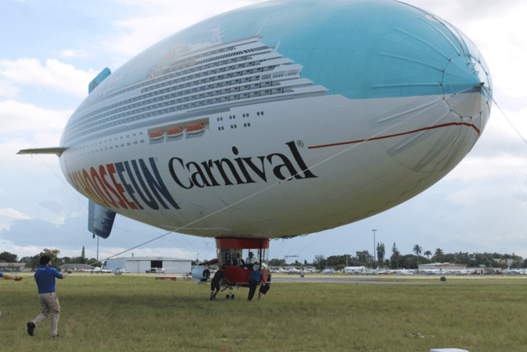 A large blimp, designed to resemble a Carnival cruise ship with the phrase "Choose Fun" written on the side, is tethered to the ground. Several people are seen holding guide ropes to stabilize the blimp, which prominently features the logo of Carnival Corporation. A small platform is suspended beneath it.