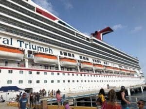 A large cruise ship named Triumph docked at a pier, with multiple lifeboats visible on the side. People are walking on the pier, some carrying bags and wearing summer clothing. The sky is clear with a few clouds. Imagine what happens if you miss the cruise ship – an adventure delayed!