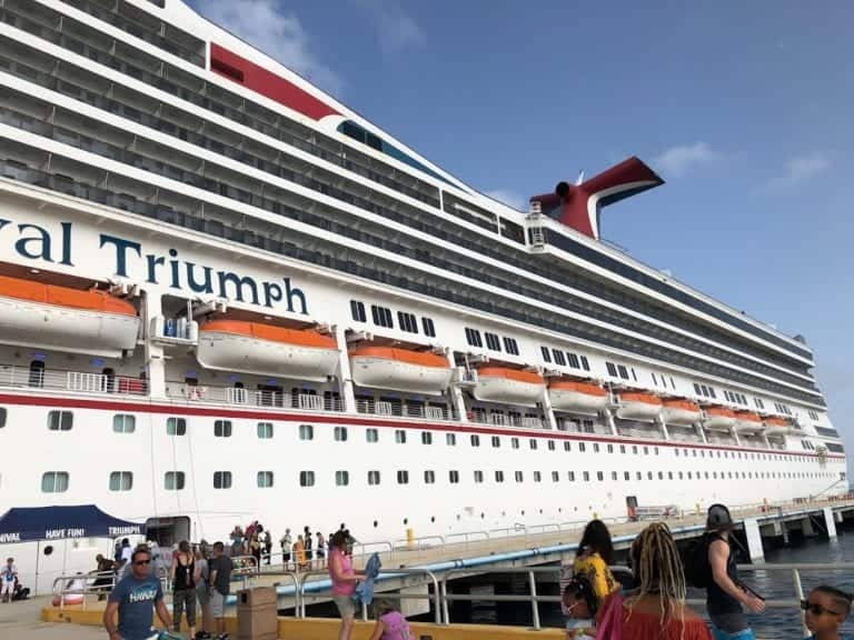 A large cruise ship named Triumph docked at a pier, with multiple lifeboats visible on the side. People are walking on the pier, some carrying bags and wearing summer clothing. The sky is clear with a few clouds. Imagine what happens if you miss the cruise ship – an adventure delayed!