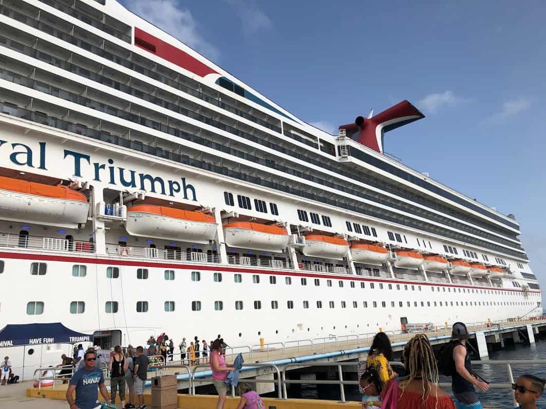 A large cruise ship named Triumph docked at a pier, with multiple lifeboats visible on the side. People are walking on the pier, some carrying bags and wearing summer clothing. The sky is clear with a few clouds. Imagine what happens if you miss the cruise ship – an adventure delayed!