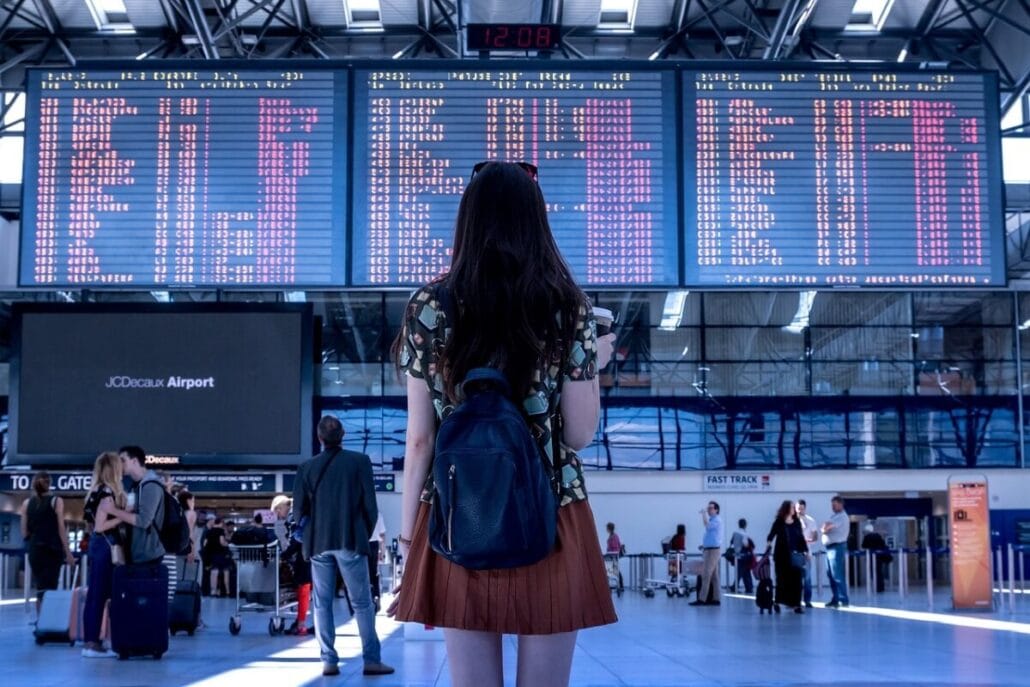 A woman with long hair and a backpack stands in an airport terminal, passport in hand, facing a large electronic flight departure board displaying various flight times and destinations. Other travelers are visible in the background.