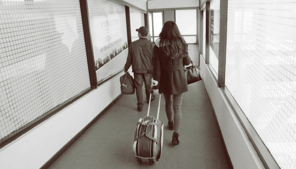 A black and white image shows two people walking down a corridor, one pulling a suitcase on wheels and the other carrying a bag. The passageway's walls and windows create a reflective, enclosed atmosphere, reminiscent of embarking on an elegant cruise or journeying with passports in hand.
