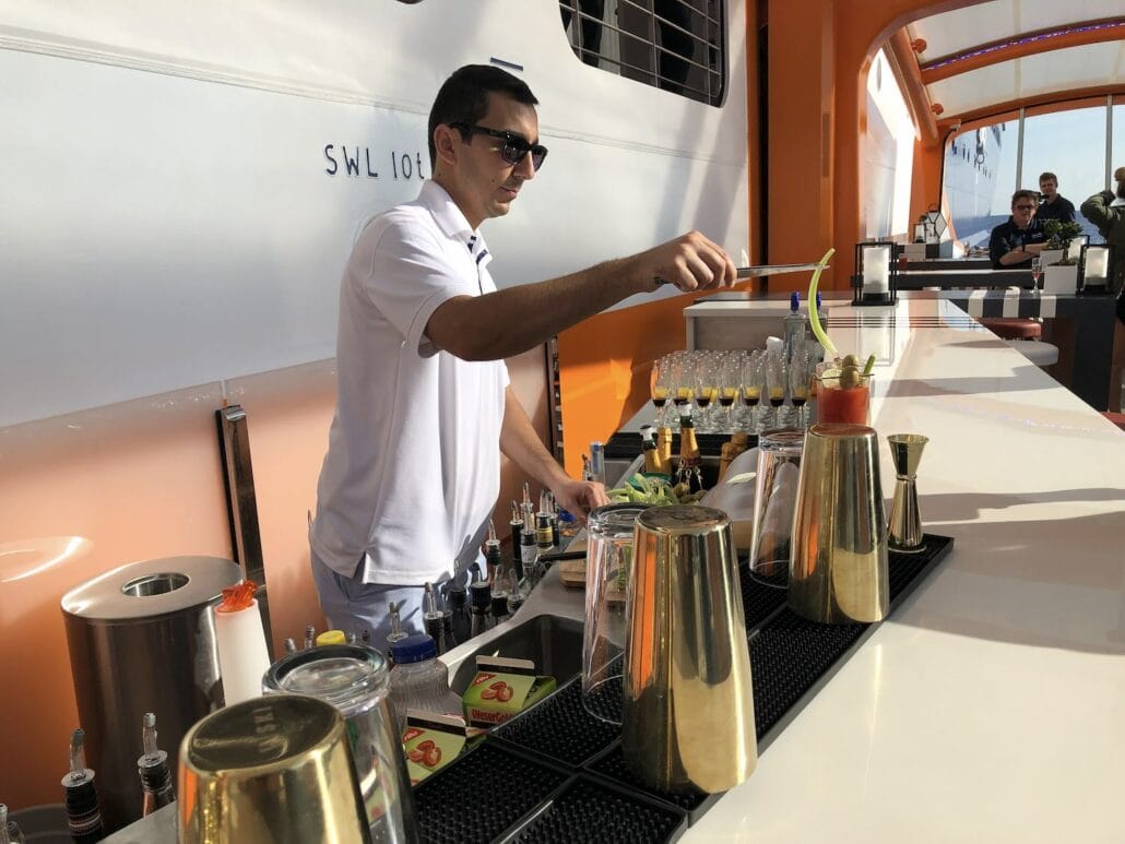 A bartender in a white uniform skillfully prepares drinks at the outdoor bar aboard the Celebrity Edge. The counter is equipped with cocktail shakers, garnishes, and glasses lined up, all against a backdrop of a white wall and a partial view of the open sky on this luxurious cruise ship.