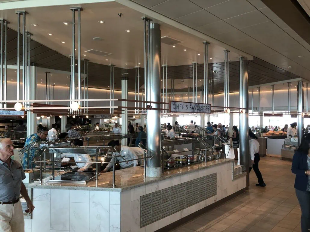 A modern buffet area aboard the Celebrity Edge cruise ship boasts a variety of food stations with people serving themselves. Silver poles and overhead lights illuminate the scene, where a sign reads "Chef's Favorites." In this delightful setting, several guests are visible in the background.