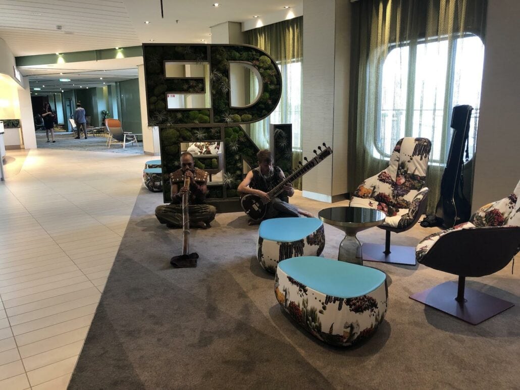In a modern lounge on the Celebrity Edge, two musicians sit on the floor playing a didgeridoo and sitar. Behind them, large letters spell out "EDEN." The space boasts patterned chairs and floor-to-ceiling windows, creating a harmonious atmosphere perfect for a relaxing cruise ship experience.