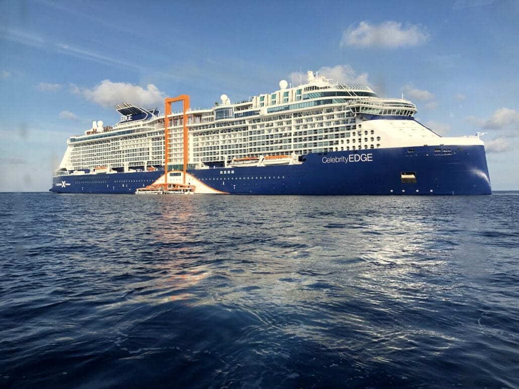 The majestic cruise ship Celebrity Edge elegantly glides over the serene ocean, its multiple decks gleaming under a clear sky. A prominent orange structure on the side adds a unique touch to this remarkable vessel, garnering glowing reviews from all who set sail aboard her.