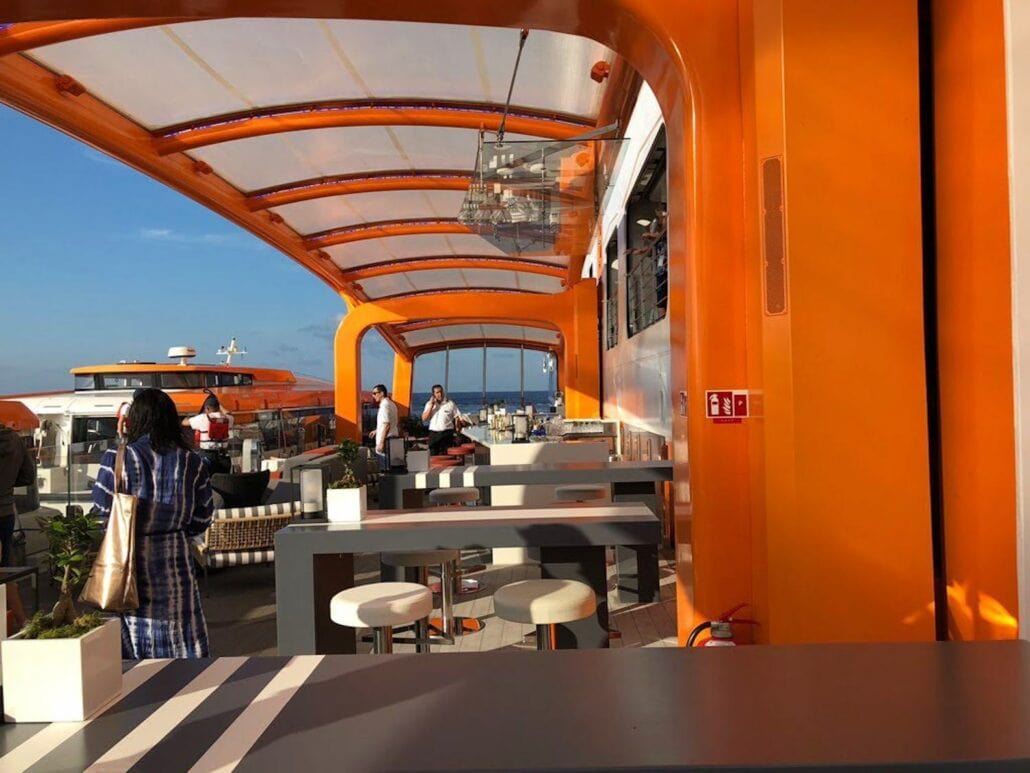 The outdoor deck of the Celebrity Edge cruise ship features bright orange structures and clear roofing. Modern bar tables and stools are arranged throughout the space, offering a chic atmosphere. A few people stand near the bar area in the background, enjoying a clear and sunny sky.