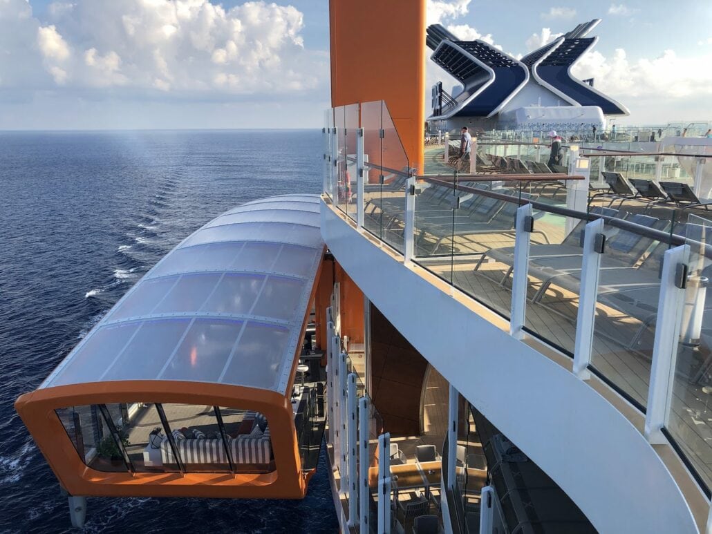 Gaze at the stunning deck of Celebrity Edge, where a sleek orange and glass lift-like structure gracefully extends over the water. With ocean views in the background, guests relax in lounge chairs, soaking up the luxury of this modern cruise ship.