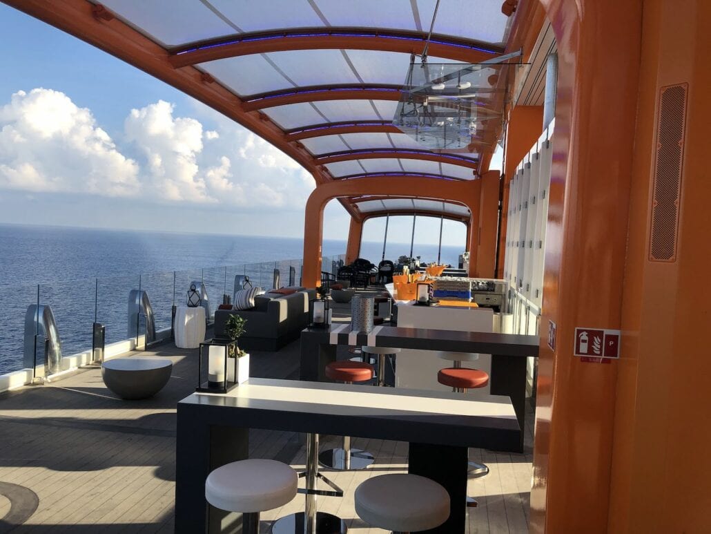 The outdoor deck of the Celebrity Edge cruise ship offers modern seating and tables under a canopy. With a partly cloudy sky, the ocean view is mesmerizing. Bar stools and decorative lamps add charm to this elegant setting, making it a perfect spot for relaxation and enjoyment at sea.