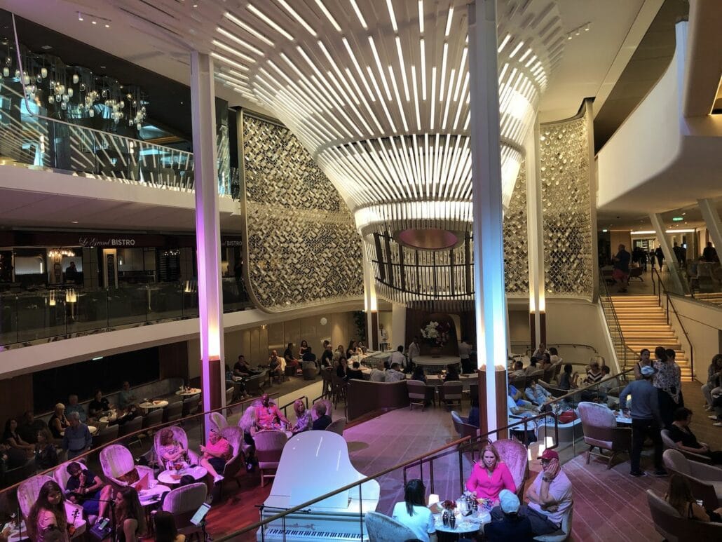 A spacious, modern lounge area aboard the Celebrity Edge offers multiple seating arrangements and a central sculpture. The ceiling features a unique, curved light installation. Guests are seated in groups, chatting and relaxing. Stairs lead to an upper level with more seating on this magnificent cruise ship.