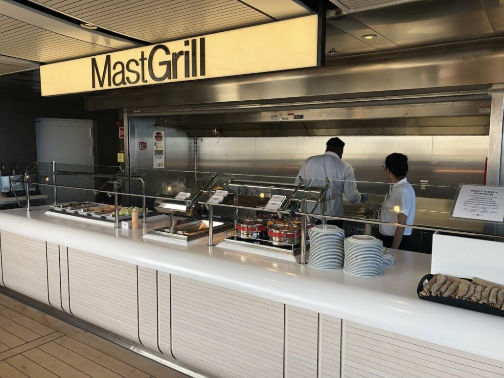 The "Mast Grill" on the Celebrity Edge showcases two chefs expertly behind the counter. With neatly displayed dishes, condiments, and stacks of plates, this grill station embodies a clean and organized dining area. A must-visit on your cruise ship adventure!