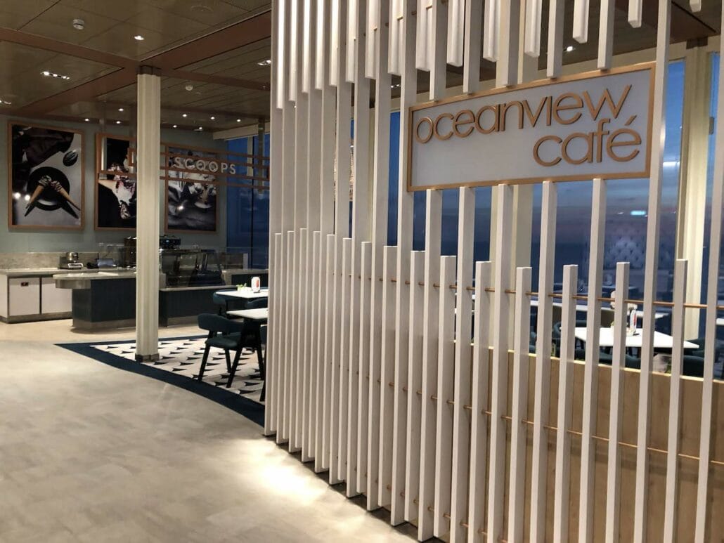 The interior of Oceanview Café on the Celebrity Edge boasts modern decor with white slatted dividers. Tables and chairs are neatly arranged, giving a pristine look to the empty café. A sign reading "Oceanview Café" is prominently displayed, inviting guests for a serene cruise ship dining experience.
