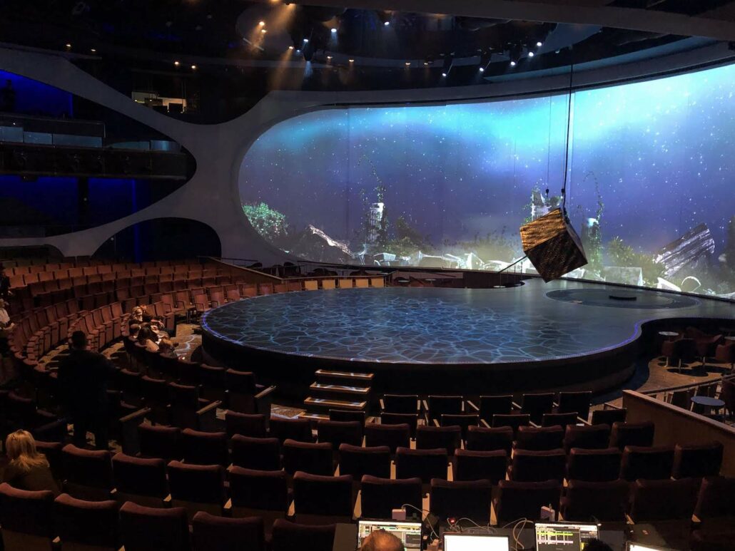 A theater aboard the Celebrity Edge features a large, circular stage with a futuristic backdrop depicting a starry sky and structures. Suspended above is a large geometric object. Rows of seats surround the stage, reminiscent of an avant-garde cruise ship show, with some people adjusting equipment in the foreground.