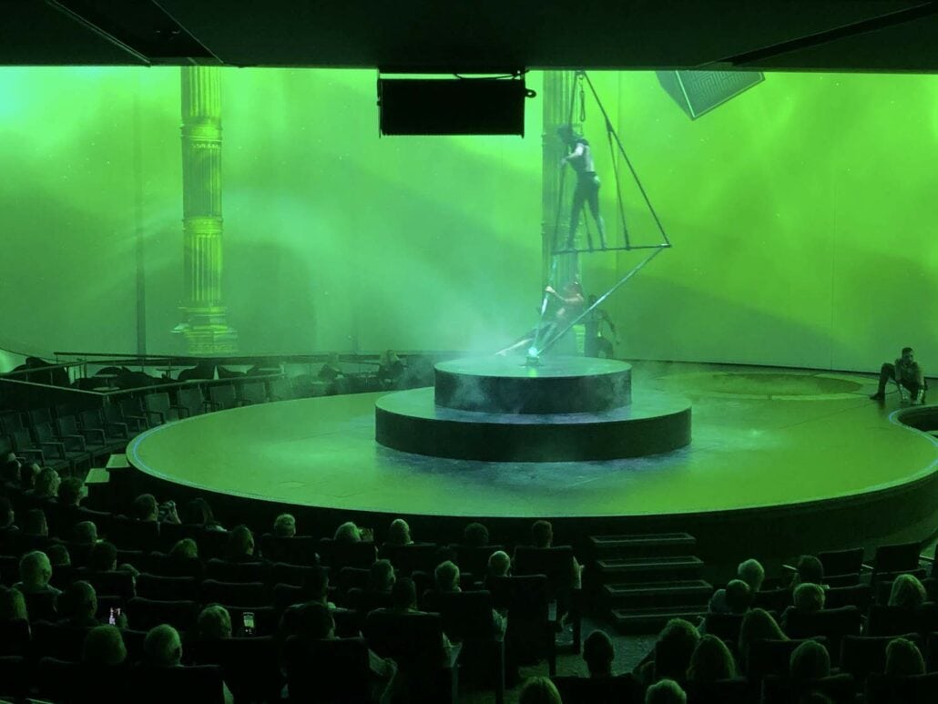 A theater performance aboard the Celebrity Edge features two acrobats on a triangular apparatus set on a round stage. The background is lit with a green hue and images of pillars, while passengers enjoy this mesmerizing show from seats surrounding the stage, making it a cruise ship highlight.