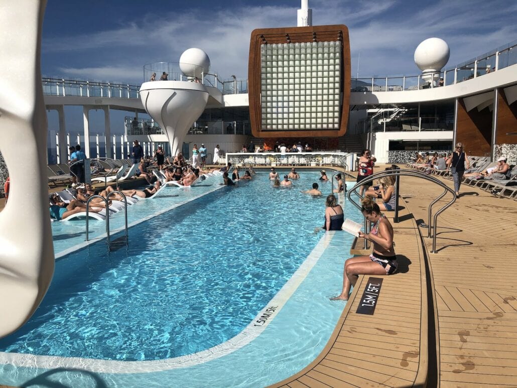 Passengers are unwinding by the pool on the stunning Celebrity Edge cruise ship deck under a partly cloudy sky. The deck's modern design includes sleek lounge chairs, a vibrant bar area, and elegant curved structures, while travelers swim, sunbathe, and socialize. 