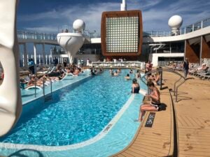 Passengers are unwinding by the pool on the stunning Celebrity Edge cruise ship deck under a partly cloudy sky. The deck's modern design includes sleek lounge chairs, a vibrant bar area, and elegant curved structures, while travelers swim, sunbathe, and socialize.
