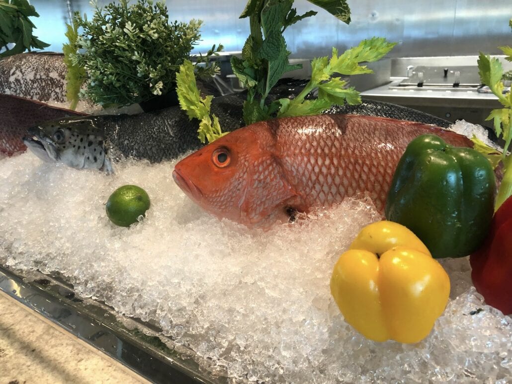 Displayed on ice with green leafy garnish, the fresh fish aboard the Celebrity Edge is surrounded by limes, green and red bell peppers, and a yellow bell pepper. Indulge in the vibrant presentation that adds to an unforgettable culinary experience on this luxurious cruise ship.