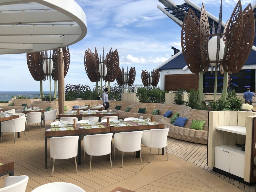 Outdoor dining area on a deck aboard Celebrity Edge, featuring white chairs and wooden tables set for a meal. Unique, leaf-shaped metal sculptures stand nearby. A curved bench has green cushions, and the ocean and sky provide a stunning backdrop to this enchanting cruise ship experience.