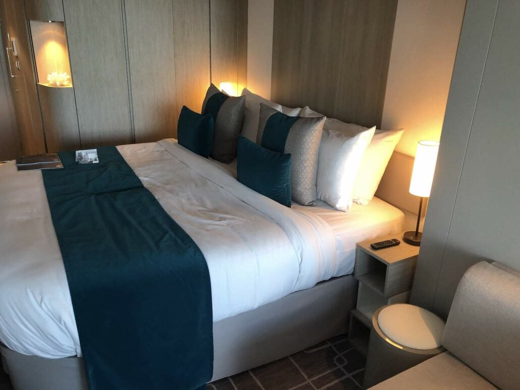 A neatly made hotel room bed with a blue throw blanket and several pillows in shades of blue and gray resembles the luxury found on a Celebrity Edge cruise ship. A nightstand holds a remote control, and a lamp casts warm light. The wood-paneled wall behind the bed features built-in shelves.
