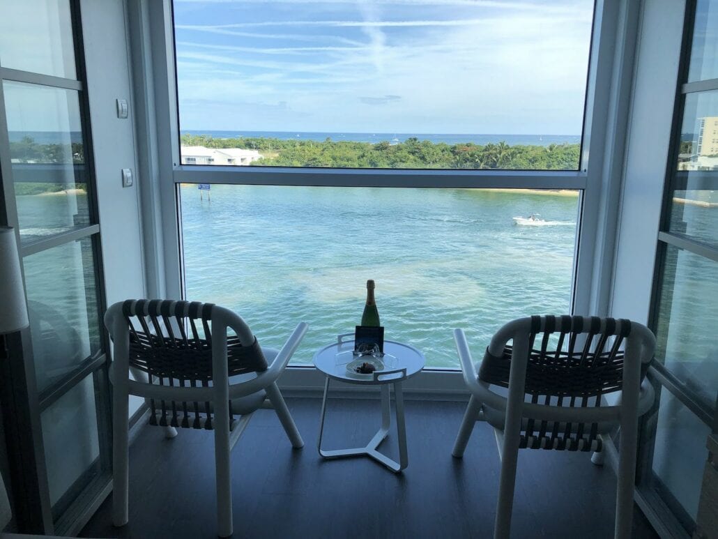 Two chairs aboard the Celebrity Edge face a floor-to-ceiling window with a view of a serene body of water and distant green trees. A small table between the chairs holds a bottle and two glasses, perfectly setting the scene. The sky is clear with streaks of clouds, offering an idyllic cruise ship experience.
