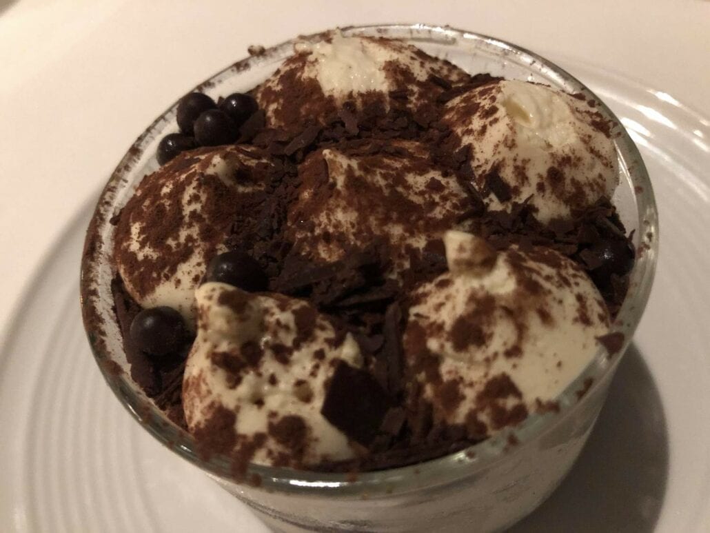 A glass bowl filled with a creamy dessert topped with cocoa powder, chocolate shavings, and small chocolate spheres—perfect for enjoying on the Celebrity Edge cruise ship as you relax and savor each indulgent bite.
