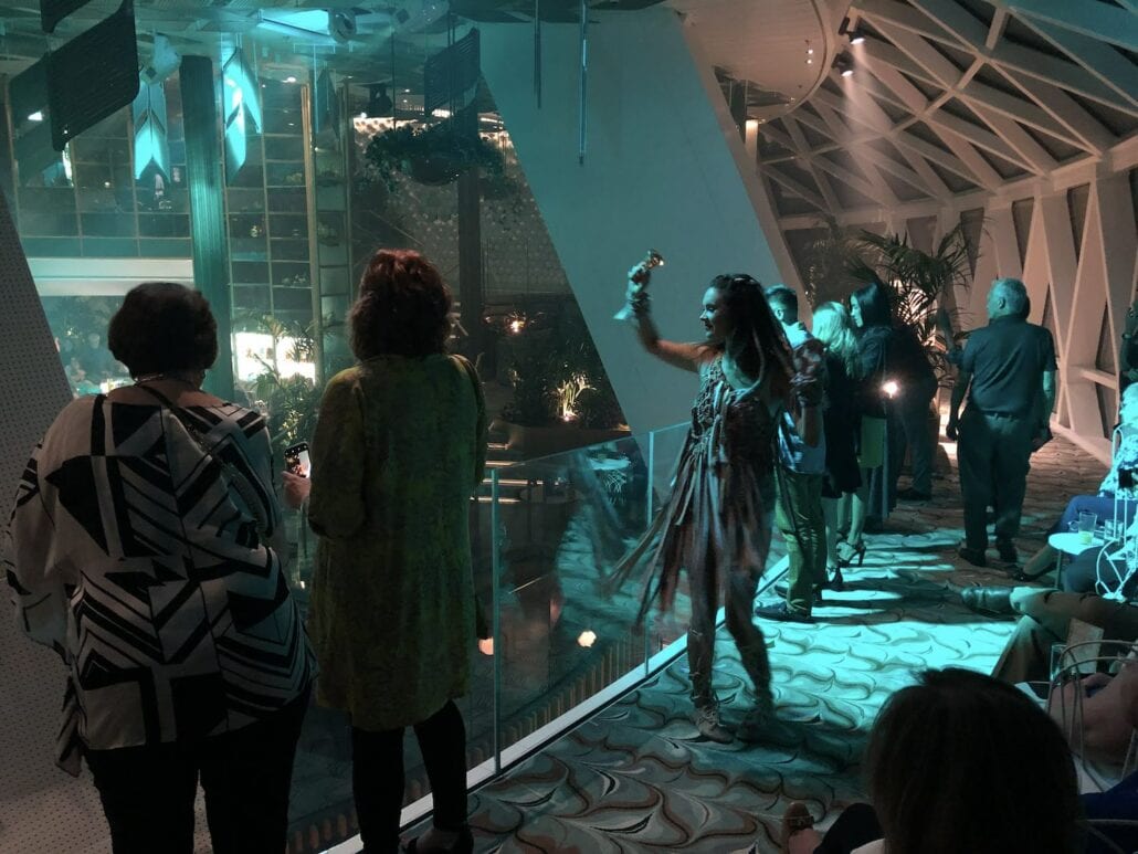 A group of people stand in a dimly lit indoor space with large windows, observing something outside. Some individuals are holding cameras. The setting, resembling the modern elegance of the Celebrity Edge cruise ship, features patterned carpet and potted plants.