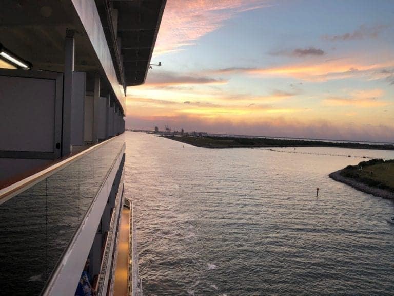 View from a Carnival Breeze cruise ship balcony looking out to a sunset over calm waters on embarkation day. The horizon shows a mix of clouds and light as the sky transitions from day to night. The ship's railing and part of the structure are visible on the left side of the image.