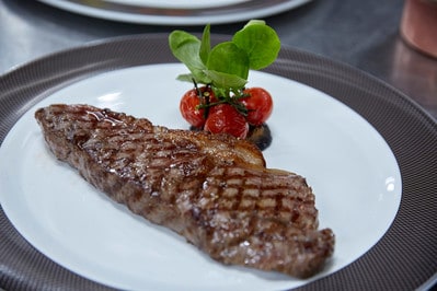 A grilled steak with distinctive grill marks sits on a white plate. Behind the steak, there are a few roasted cherry tomatoes and a garnish of green leaves. The dish, part of the cruise line's expanded steakhouse menu, is placed on a dark woven placemat.