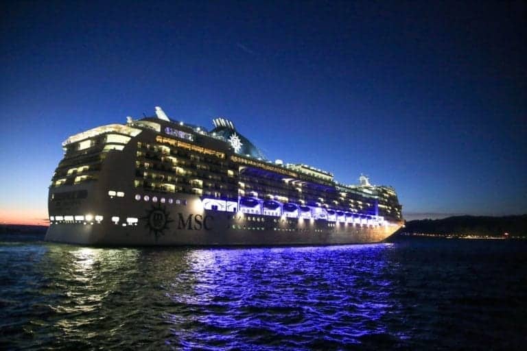 A large cruise ship with the letters "MSC" on the side is illuminated with blue and white lights as it sails on the water during the evening. The sky is darkening, and the ship's reflection is visible on the water's surface, perfect for anyone looking to tick off a bucket-list cruise experience.