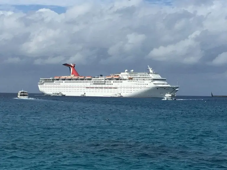 A large white cruise ship with a distinctive red funnel is sailing on a calm ocean under a partly cloudy sky. Several smaller boats surround it in the water. The backdrop features an expansive horizon.