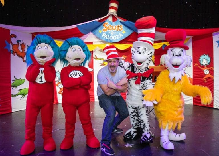 A group of five people, including Joey Fatone and four individuals in Dr. Seuss character costumes, pose in front of a Carnival's Seuss-a-Palooza sign. The characters include Thing 1, Thing 2, Cat in the Hat, and The Lorax. The background displays a colorful, Dr. Seuss-themed setup.