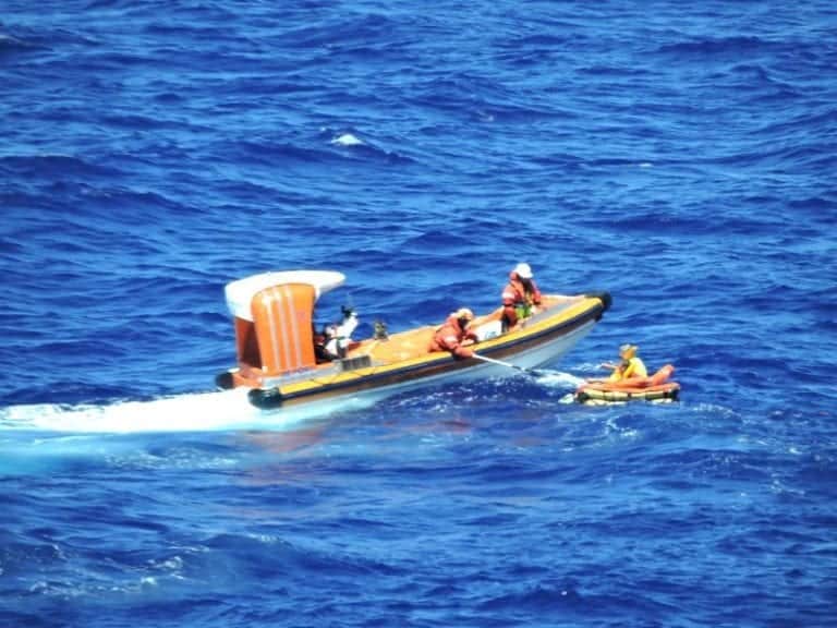 Four people in orange gear, part of a cruise ship crew, are on a motorized rescue boat in the middle of the ocean. The boat is assisting a person in a small inflatable raft. The blue sea surrounds them.