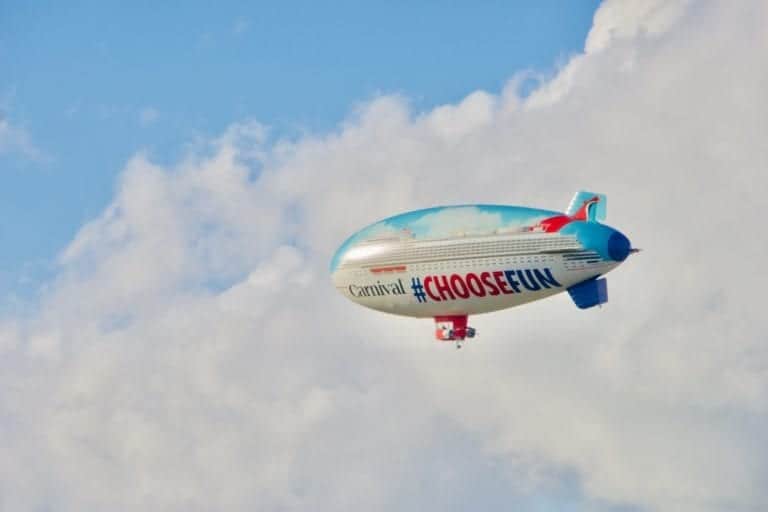 A blimp floats in the sky with "Carnival" and "#CHOOSEFUN" written on its side against a backdrop of clouds, promoting Carnival Cruise Line and its adventures by sea and by air.