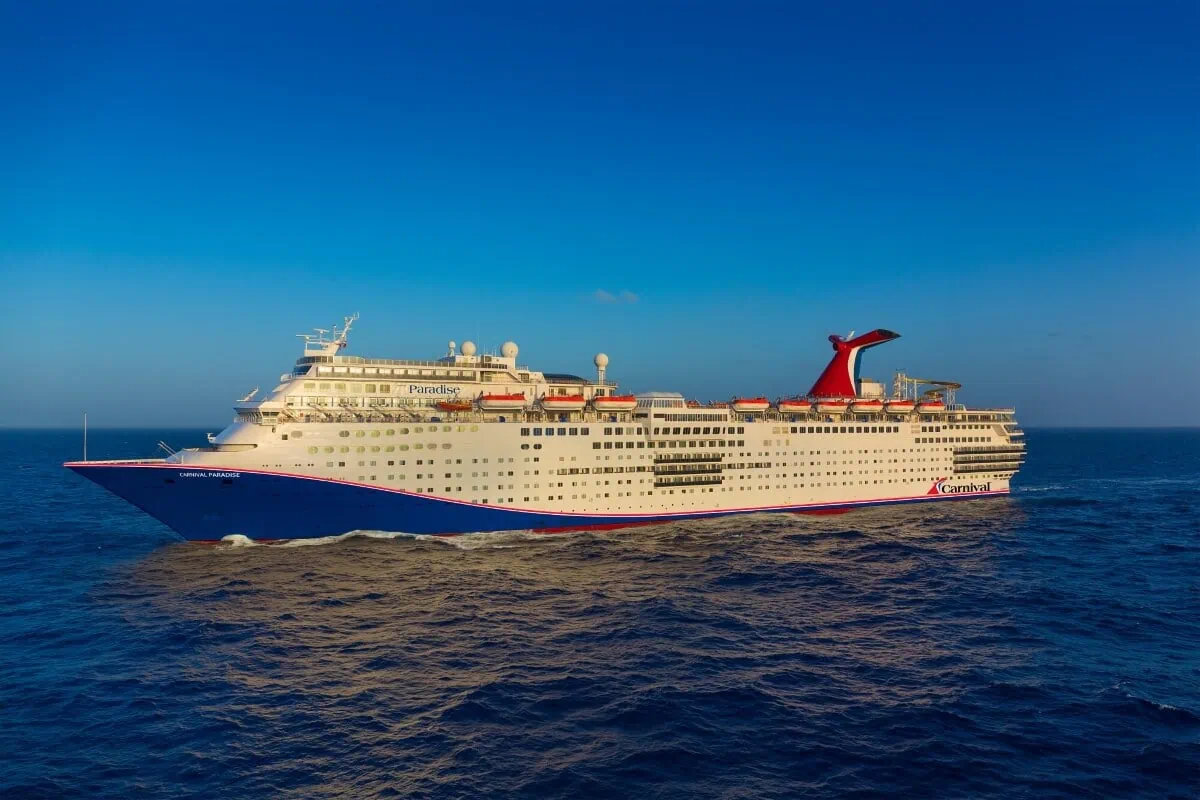 A large Carnival Cruise ship is sailing on the open sea under a clear blue sky. The vessel is white with red and blue accents and several decks, featuring lifeboats and a distinctive red funnel.